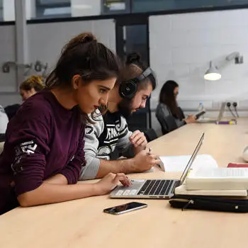 female student working on her laptop in the university library with other students in the background