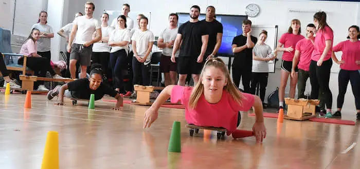 female students on a body board race across the floor while a group of other students watch