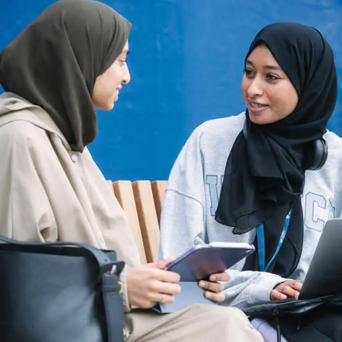 Two women in hijabs sit side by side on a bench, one holding a tablet while the other works on a laptop