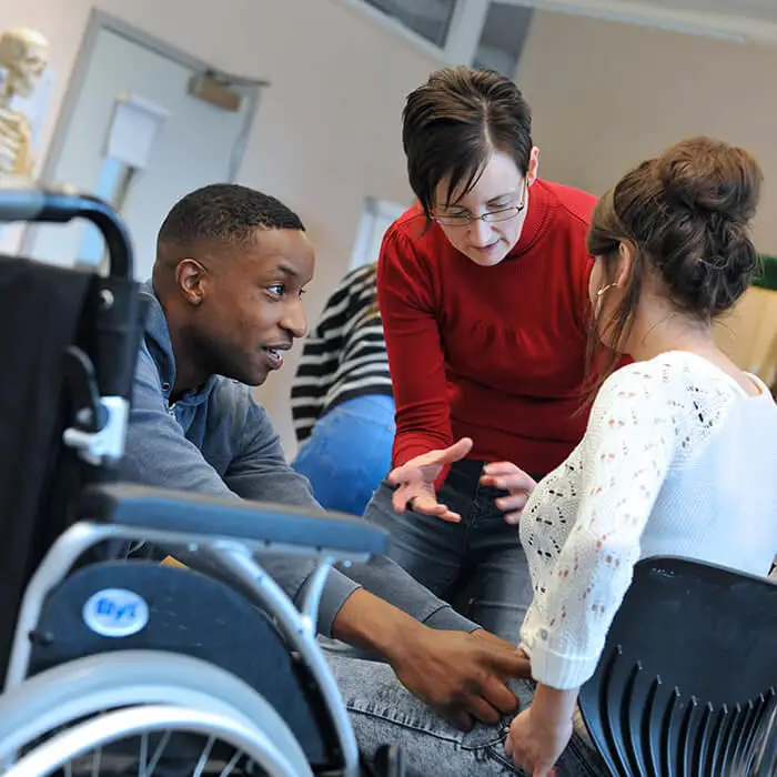 female lecturer explaining occupational therapy practical exercise to two students with a wheelchair in the front