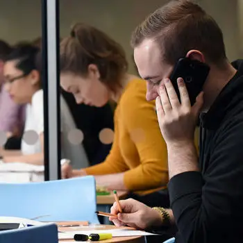 male student taking a phone call while studying in the university library with other students in the background