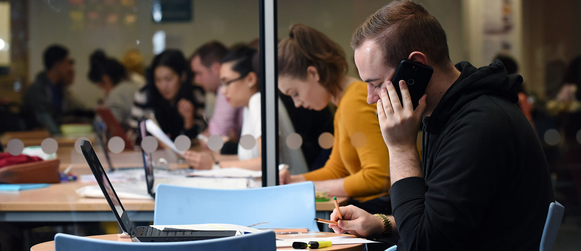 male student taking a phone call while studying in the university library with other students in the background