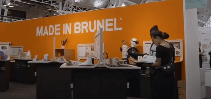 A vibrant orange trade booth displays the phrase MADE IN BRUNEL, featuring product showcases and attendees engaging with the display
