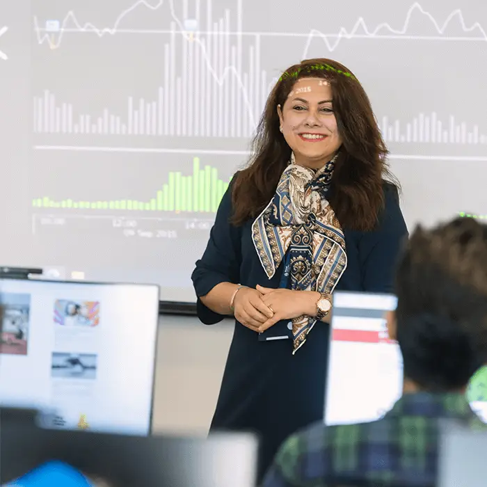 A woman stands in front of a large screen displaying financial data while presenting to an audience seated at desks with computers