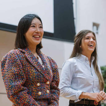 Two women standing indoors, one wearing a colorful textured jacket with pearl buttons, the other in a white button-up shirt