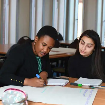 Two female students sitting at a desk and using markers to write on A3 paper