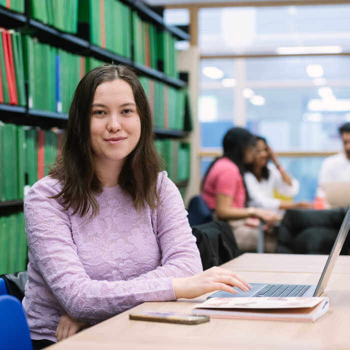 student with a laptop and phone at a desk in a library