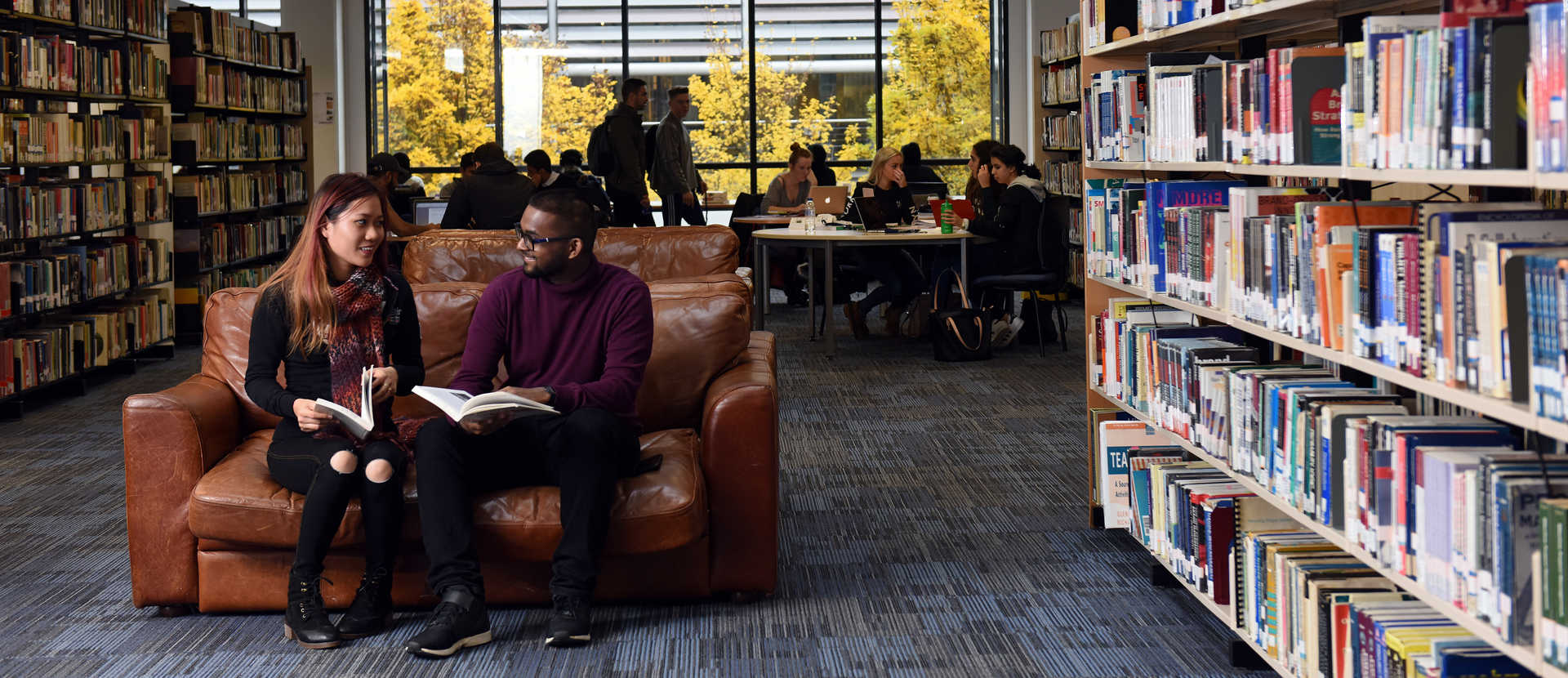 Two students reading book on a brown leather sofa in a library