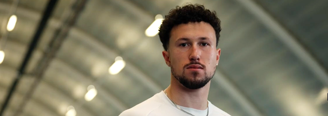 Man with curly hair and beard wearing a white sports shirt with a British flag and a red Nike logo indoors.
