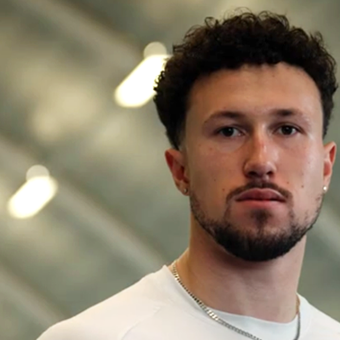Man with curly hair and beard wearing a white sports shirt with a British flag and a red Nike logo indoors.