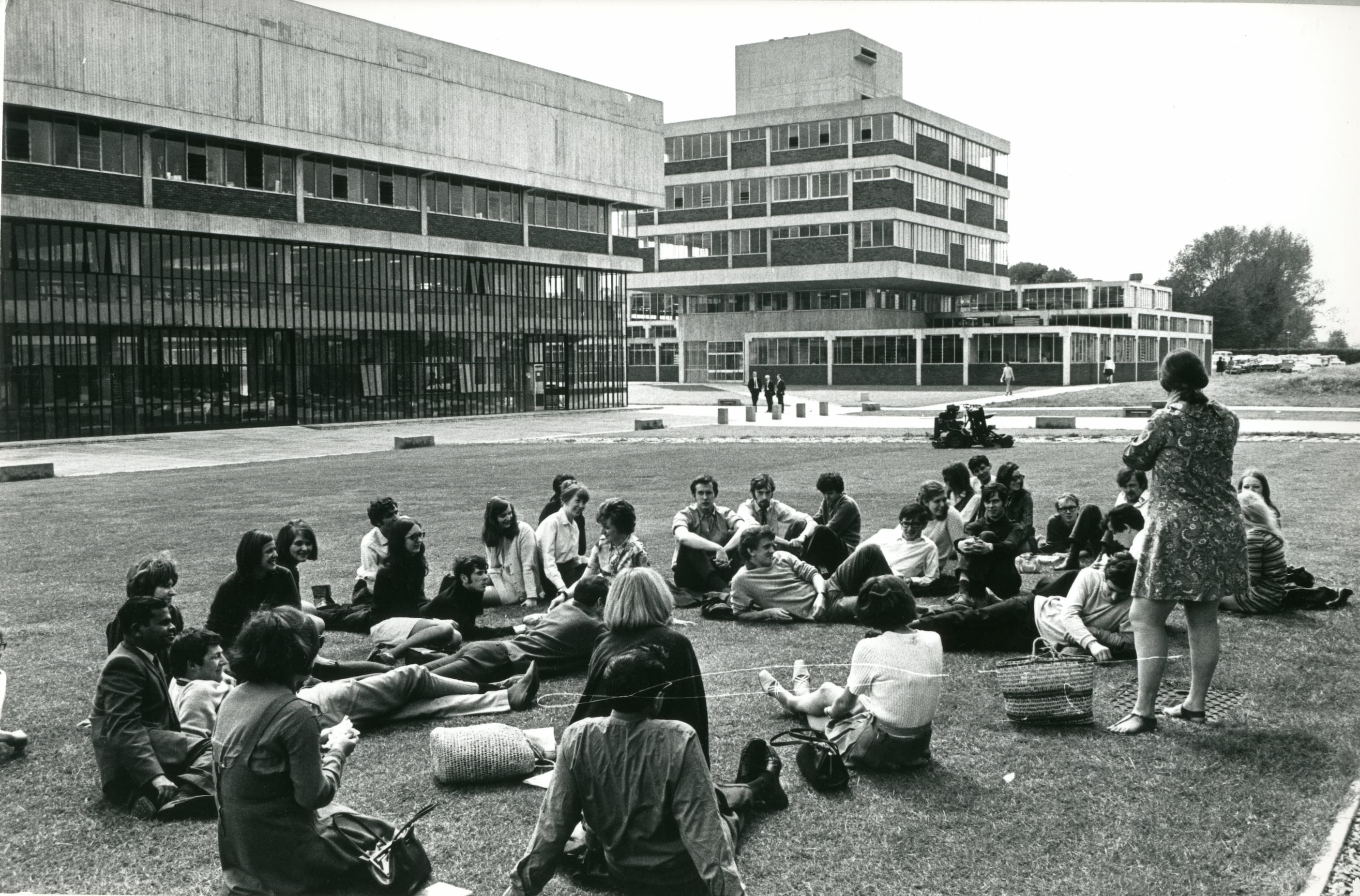 Black and white image of students sitting on grass in front of buildings 