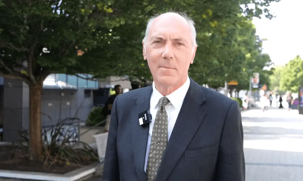 Man in a dark suit and tie stands outdoors on a sunny sidewalk with trees and buildings in the background
