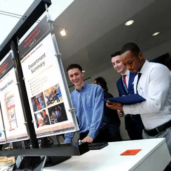 A group of students and professionals engaged in a discussion at a technology display, examining posters on computer science projects