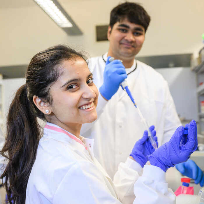 Two scientists in lab coats and gloves working with a pipette in a laboratory, smiling at the camera.