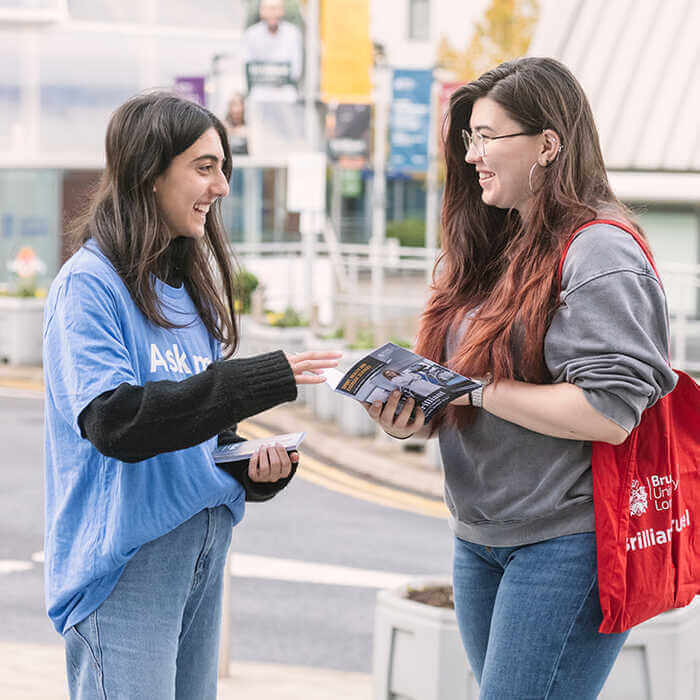 Two young women smiling and talking, one handing a brochure to the other outdoors near a street