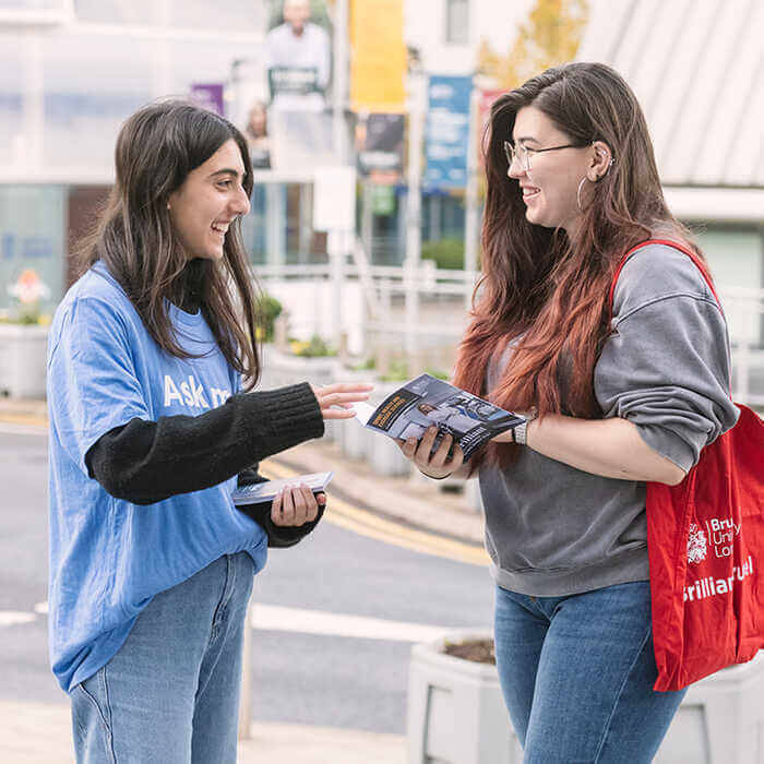 Two young women smiling and talking, one handing a brochure to the other outdoors near a street