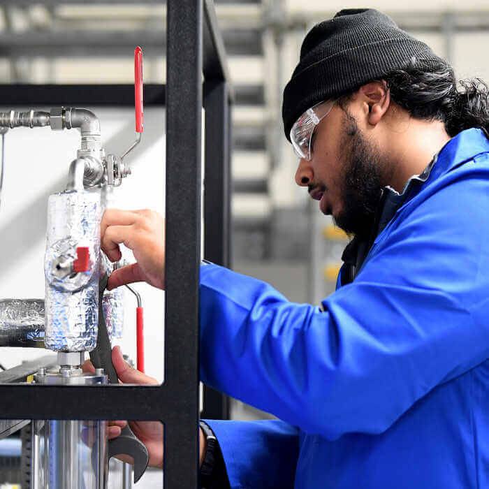 Technician in blue coat and safety glasses adjusting industrial valve with red handles in a factory setting