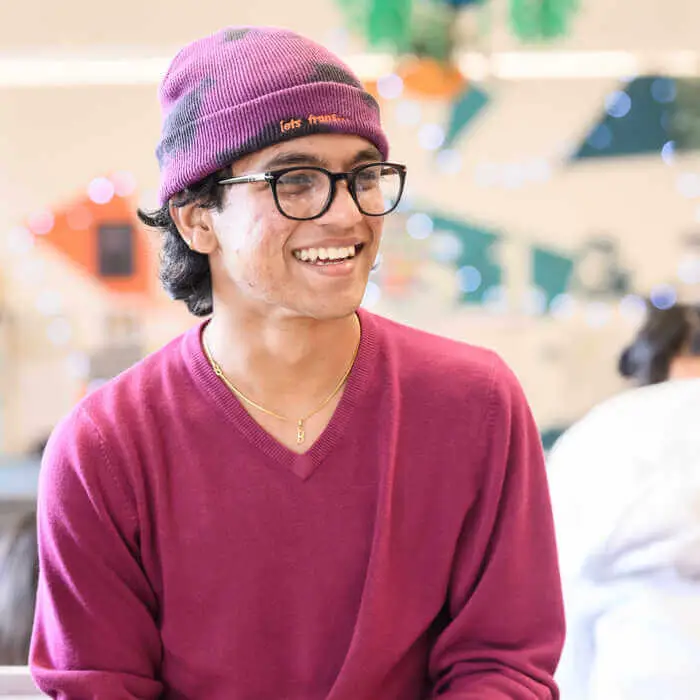 Young man wearing glasses, a purple beanie, and a matching sweater smiling in a brightly lit indoor setting