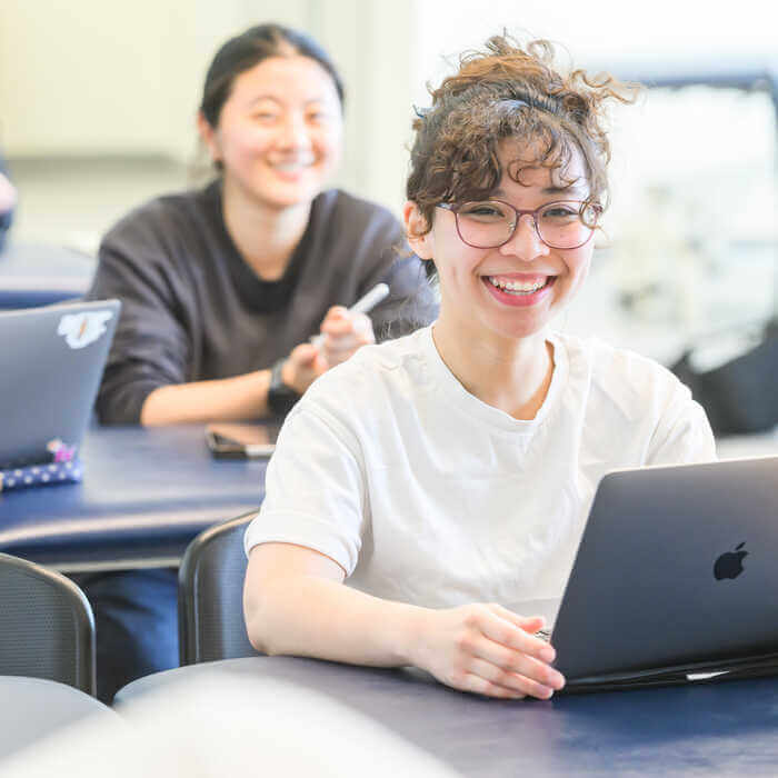 Two students smiling while sitting at desks with laptops, one in a white shirt and glasses