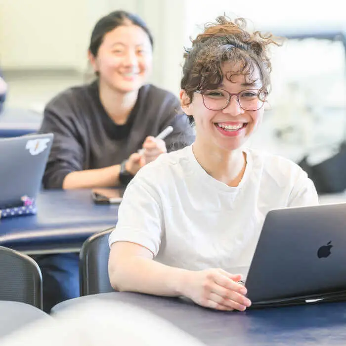 Two students smiling while sitting at desks with laptops, one in a white shirt and glasses