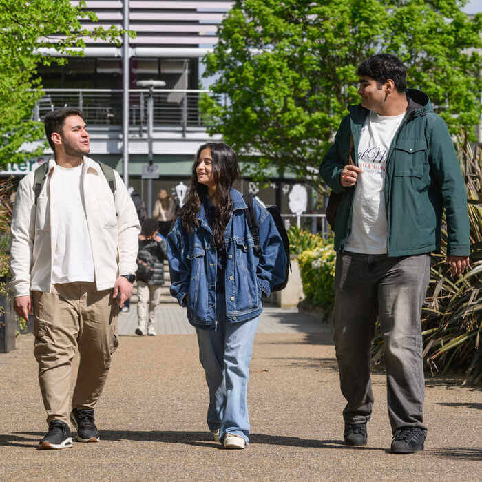 Three students with backpacks walk and chat on a sunny urban path lined with greenery and modern buildings.