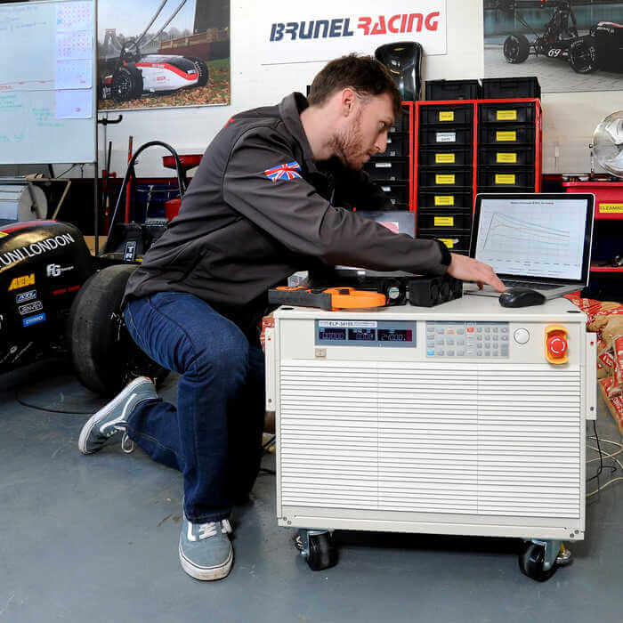 Man working on a laptop on top of a large machine in a garage with racing posters and a race car nearby