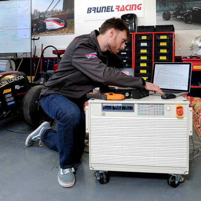 Man working on a laptop on top of a large machine in a garage with racing posters and a race car nearby