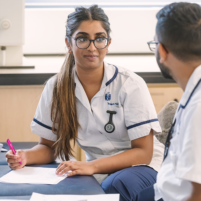 Two nursing students in uniform discussing notes in a classroom, one holding a pink pen and wearing glasses.