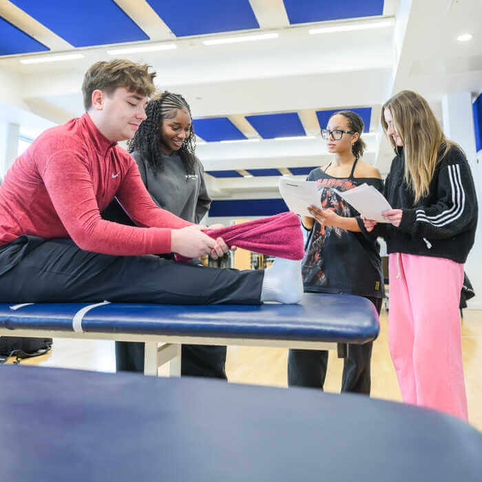 Four young people in a gym setting, one sitting and stretching leg, others observing and holding papers.