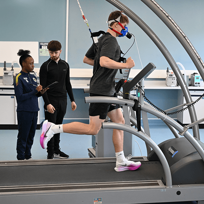 Person running on treadmill wearing a respiratory mask, monitored by two observers taking notes in a lab setting.