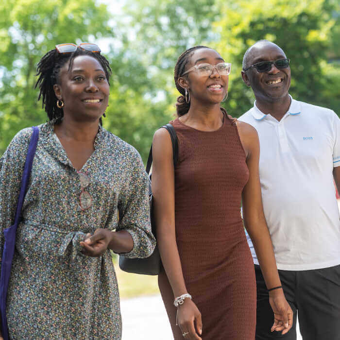 Three smiling people, two women and one man, walk outdoors on a sunny day with trees in the background