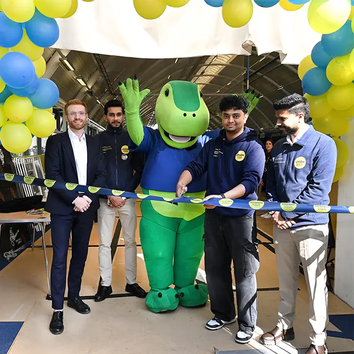 Five people, including a green dinosaur mascot, cut a University of London ribbon under blue and yellow balloon arch.