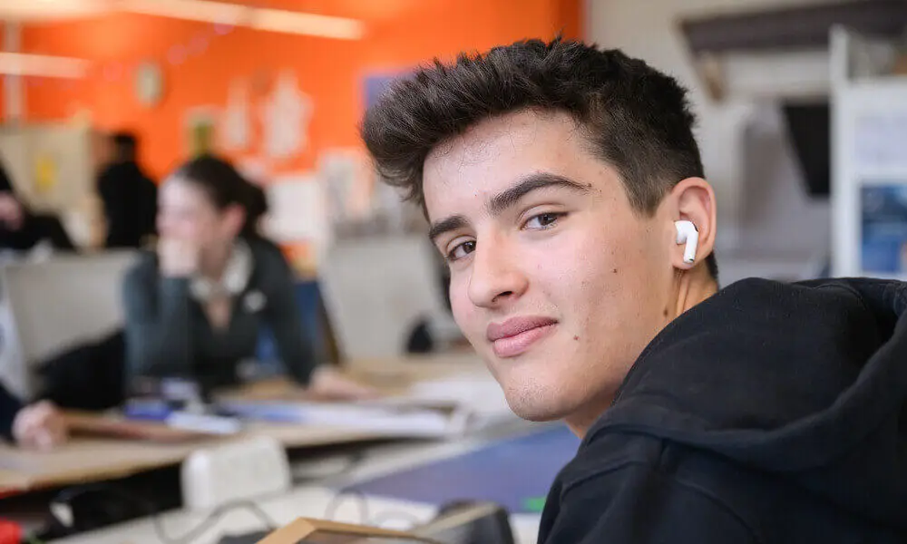 Student wearing wireless earbuds and a black hoodie sitting at a desk in a classroom
