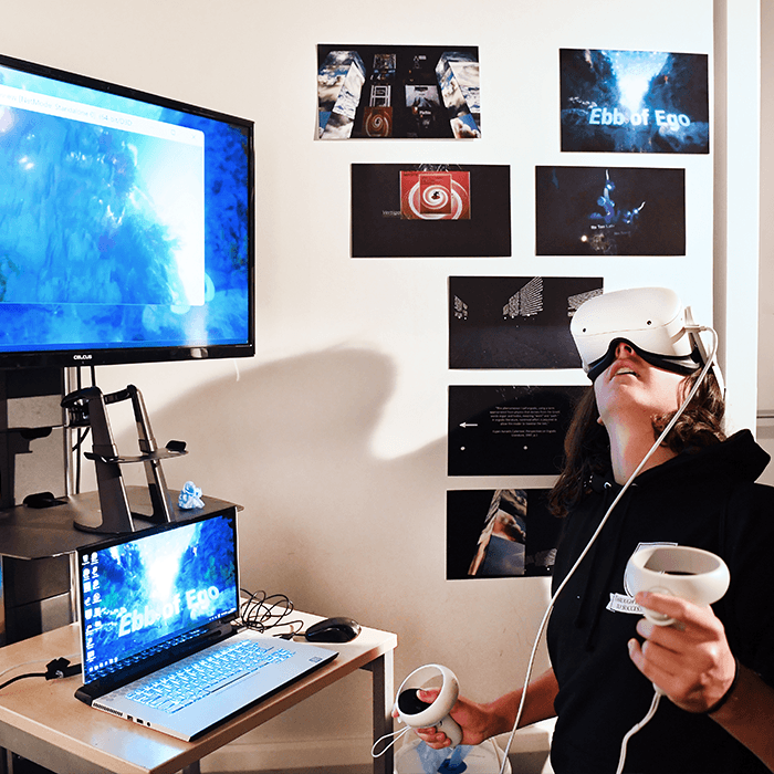Person using a VR headset and controllers in a room with a laptop and monitor