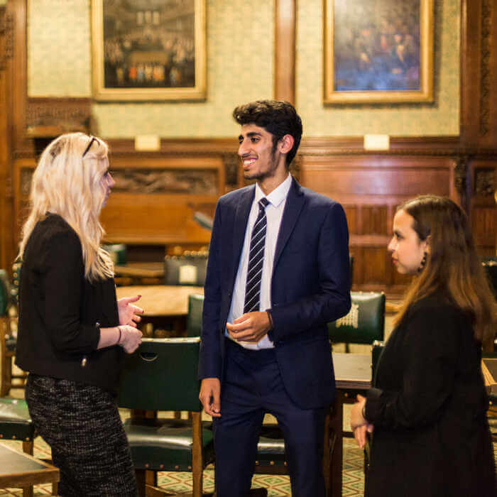 Three people in formal attire engaged in conversation in a room with wood paneling and framed paintings