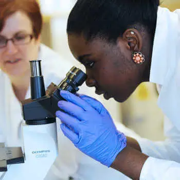 female student in white coat looking into a microscope in a science laboratory