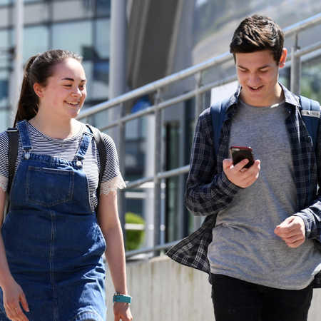 female and male student walking across campus, one of them is looking at his phone