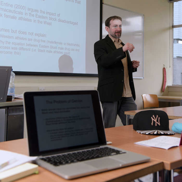 lecturer talking on front a projector display and a laptop in the foreground