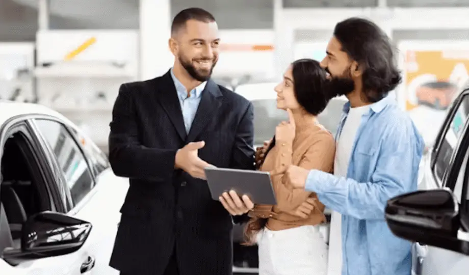 A-car-salesman-discussing-options-with-a-couple-in-a-dealership-holding-a-tablet- surrounded-by-white-cars-thumb