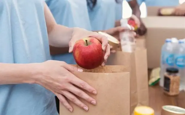 Volunteers in blue shirts packing groceries, placing a red apple into a brown paper bag on a table