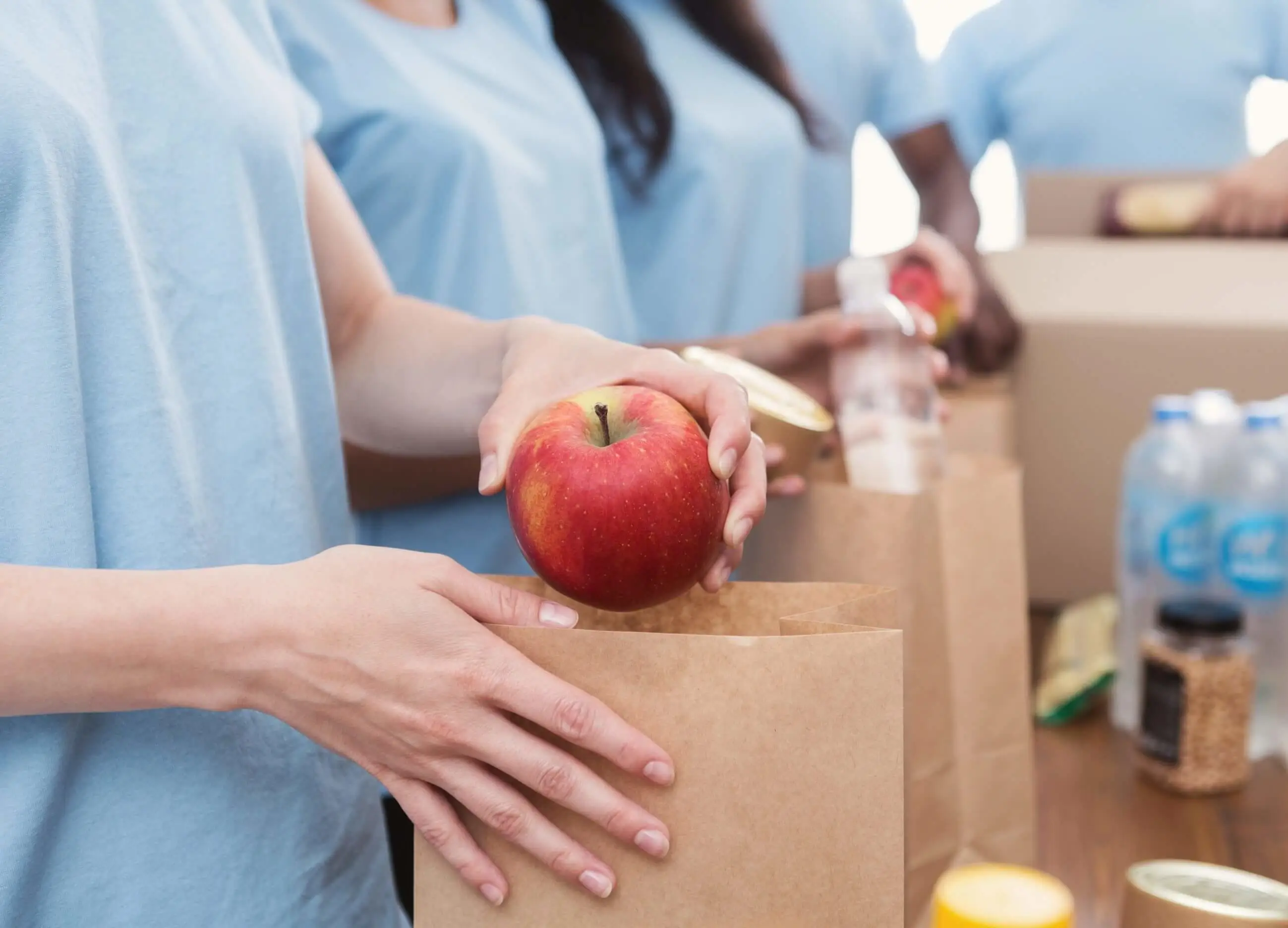 Volunteers in blue shirts packing groceries, placing a red apple into a brown paper bag on a table
