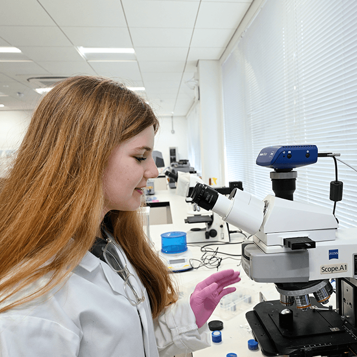 A person in a lab coat and pink gloves interacts with a microscope in a modern laboratory setting, surrounded by scientific equipment