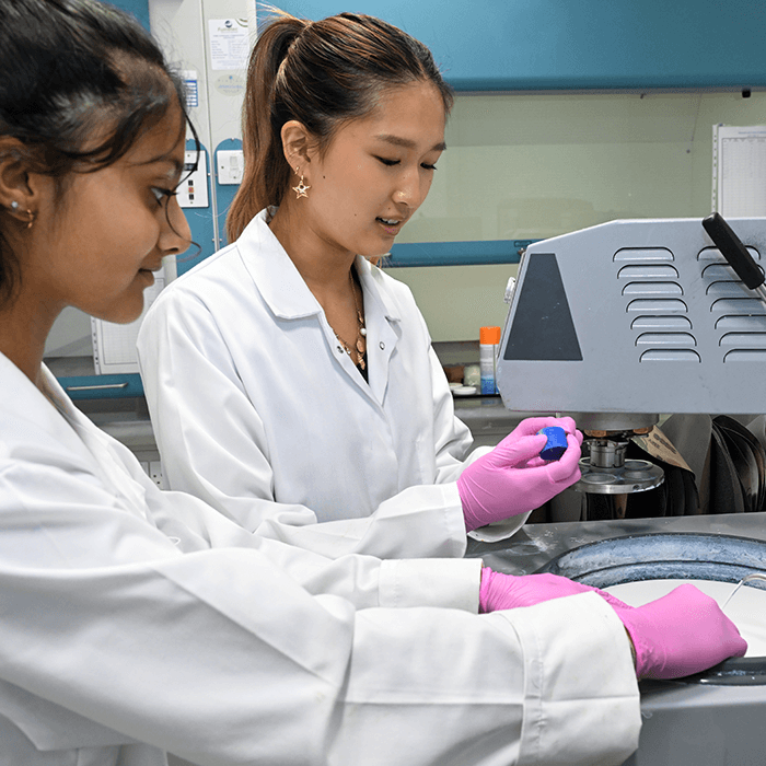 Two laboratory workers in white lab coats and purple gloves using a machine, handling a blue object and observing the process