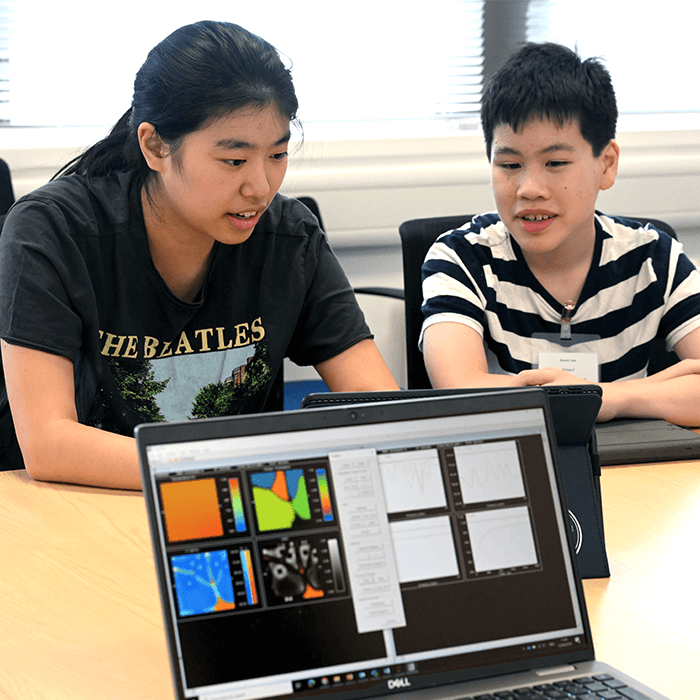 Two students are seated at a table, focused on a computer displaying colorful data visualizations and graphs