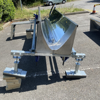 A polished, metal trough mounted on supports, resting on a parking lot with greenery and a parked car in the background.