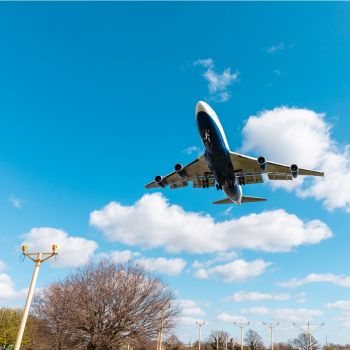 A large airplane flying overhead preparing to land on Heathrow Airport with trees and lamps visible below.