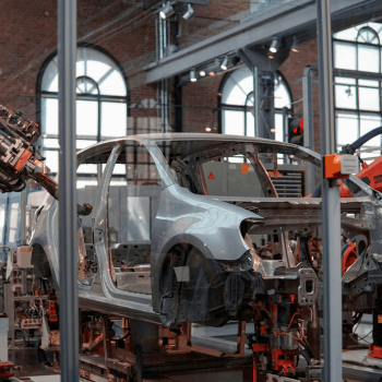 An industrial setting featuring a robotic arm assembling a silver car chassis on a production line with large windows in the background.