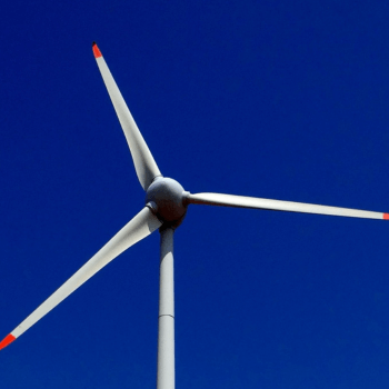 A tall wind turbine against a clear blue sky, featuring three large white blades with red tips.