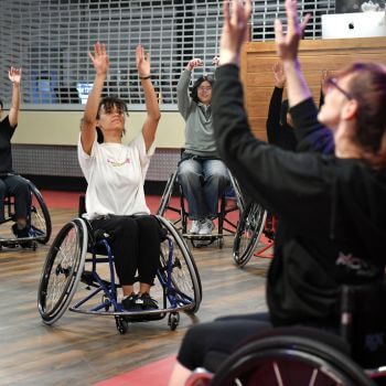 A group of individuals in wheelchairs participate in an engaging activity, raising their arms.