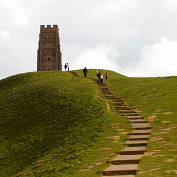 People ascending steps on a hill towards a stone tower called Glastonbury Tor under a cloudy sky.